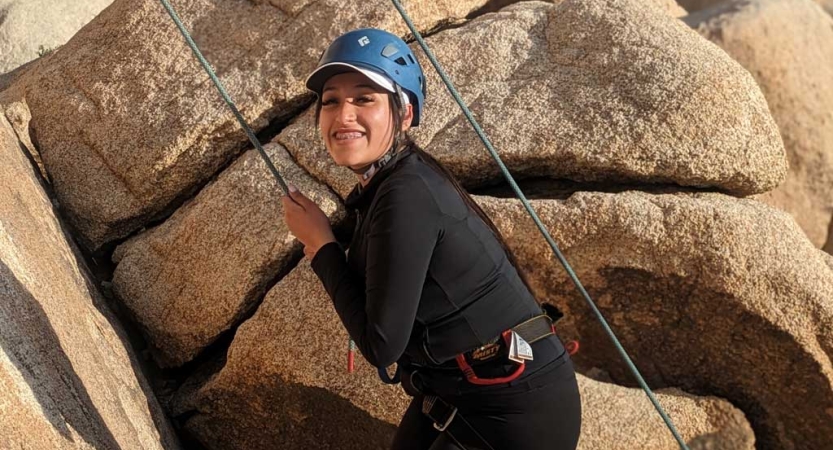 A person wearing safety gear is secured by ropes as they rock climb. They are smiling at the camera. 